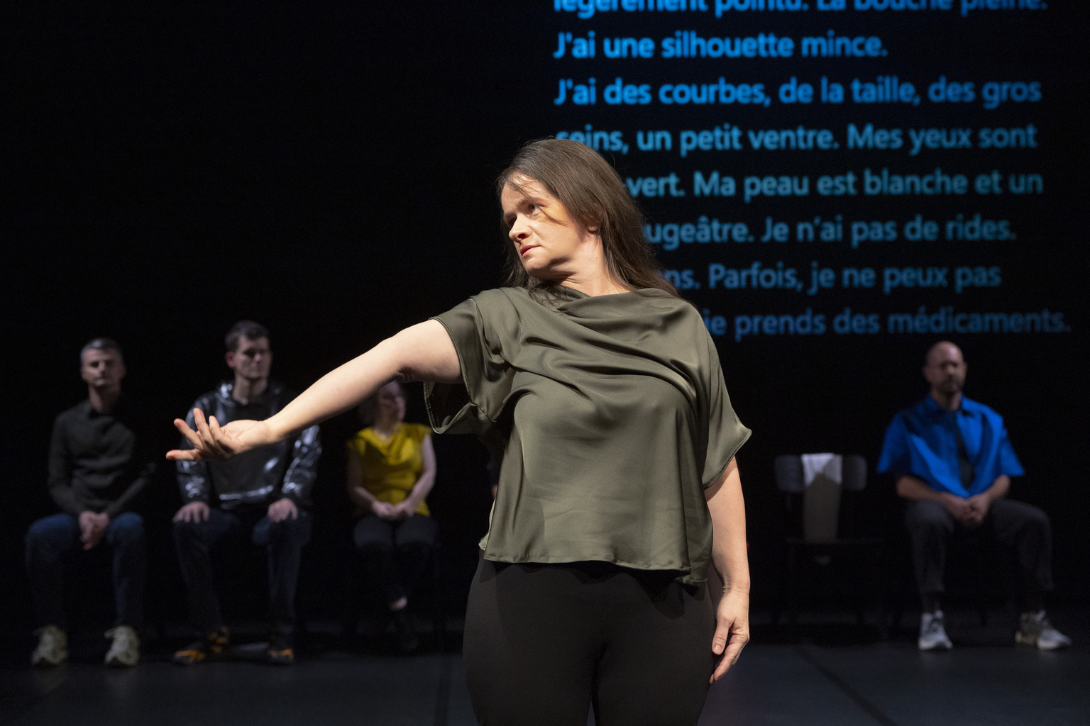 Dis Dis Contact performance - photo by Laurent Philippe. The image shows Snježana Bulatović standing in the foreground, extending her arm with a graceful gesture. She is wearing a loose-fitting, silky green top. Her expression appears focused and intent. Behind her, four other seated performers are partially visible, sitting against a dark background. A large text in French is projected on the backdrop, as a part of a performance containing Snježana´s self-description. The scene is theatrical, with dim lighting focusing on Snježana, highlighting her movement and presence on stage.